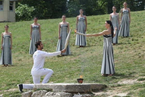 扮演最高女祭司的演員瑪麗·米娜（前右）點(diǎn)燃第一棒火炬手——希臘運(yùn)動(dòng)員、東京奧運(yùn)會(huì)男子賽艇項(xiàng)目金牌得主斯特凡諾斯·恩圖斯科斯手中的火炬。新華社記者 趙丁喆 攝